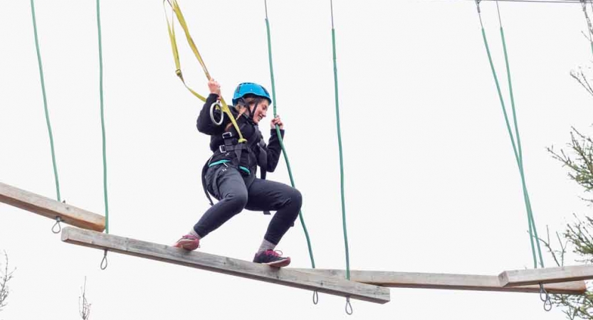 A person wearing safety gear and secured by ropes balances on an obstacle on a high ropes course. 